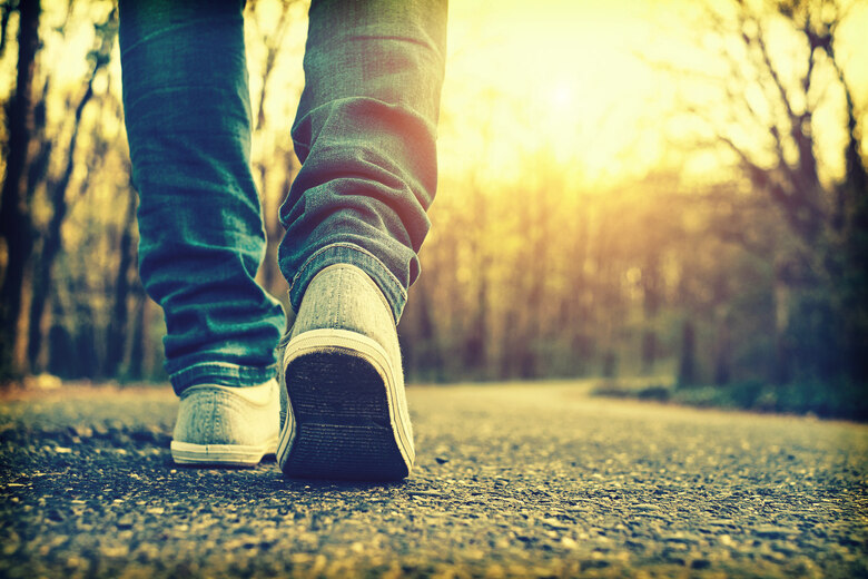 Man sieht die Beine eines Menschen. Er geht dem Sonnenlicht auf einem geteerten Waldweg entgegen.